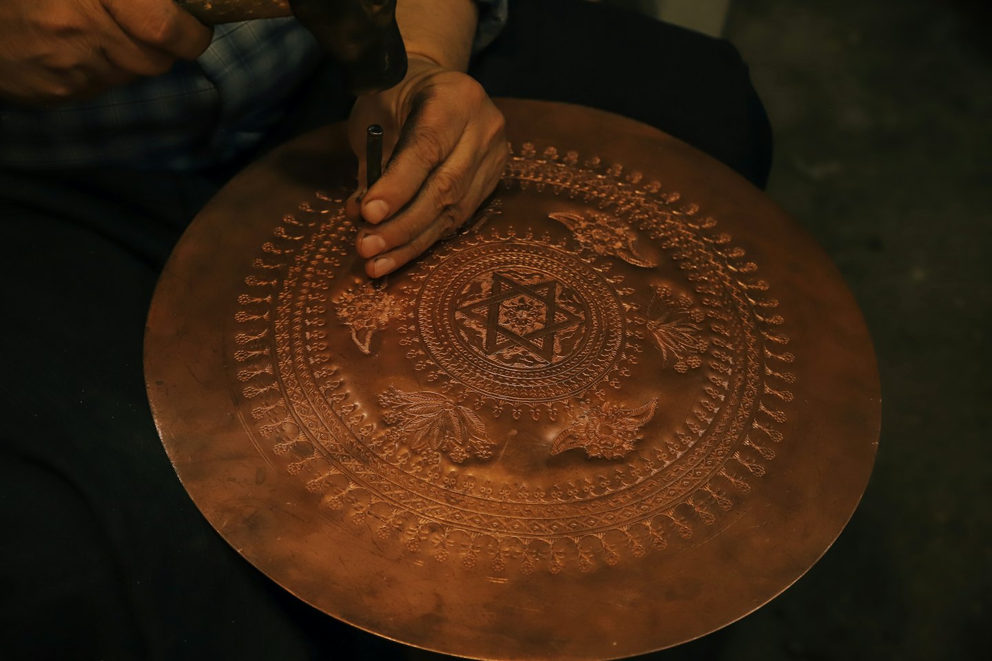 Hands use a hammer and punch to emboss a copper plate.